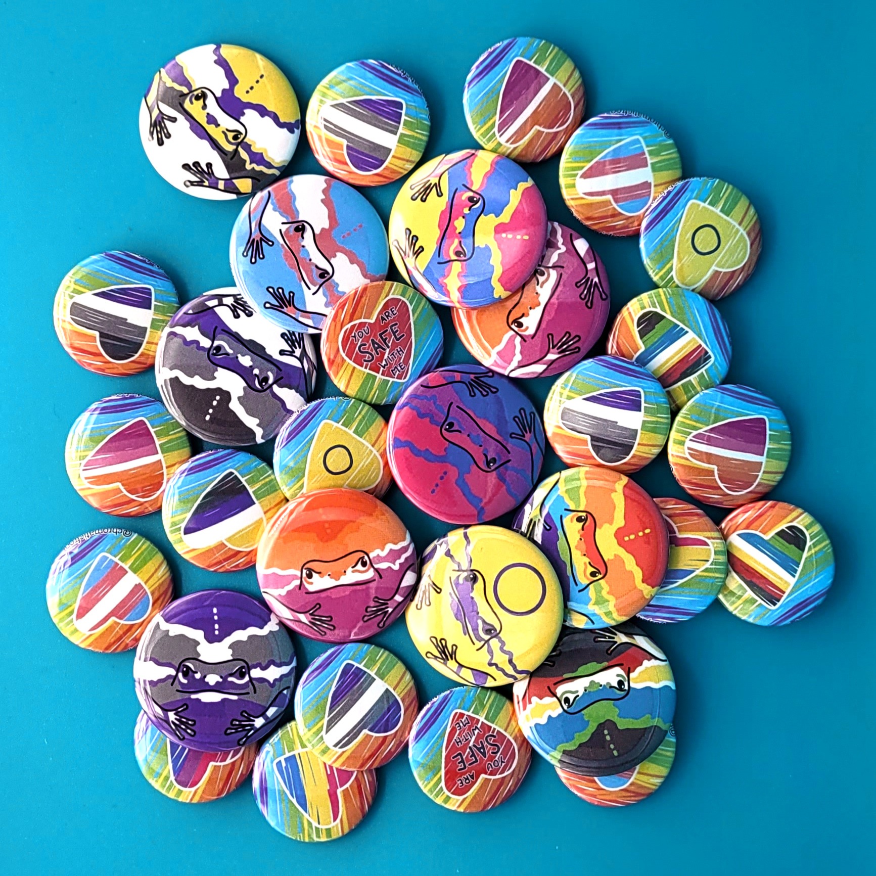A jumble of pinback buttons in various sizes featuring frogs and hearts in various pride flag colours.